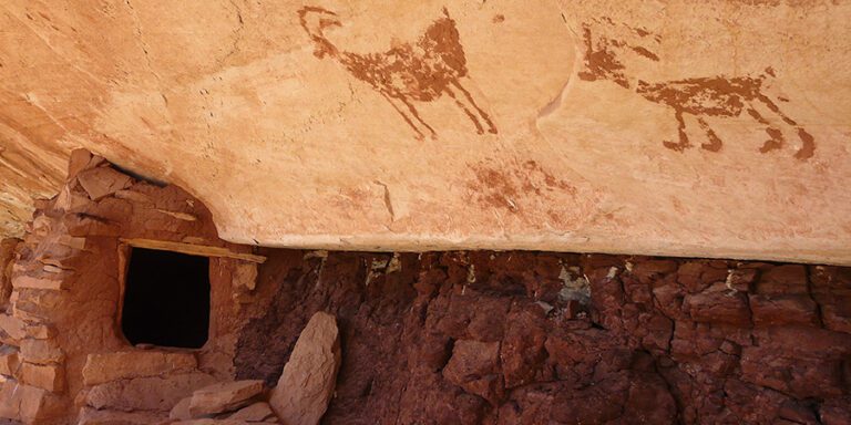 Cliff dwelling with a small dark entrance, red stone walls, and two faded animal petroglyphs on the ceiling above.