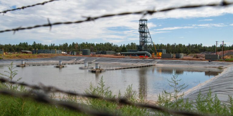 A large lined industrial pond with aeration equipment in the foreground, viewed through barbed wire, with a mine headframe in the background, Pinyon Plain uranium mine.